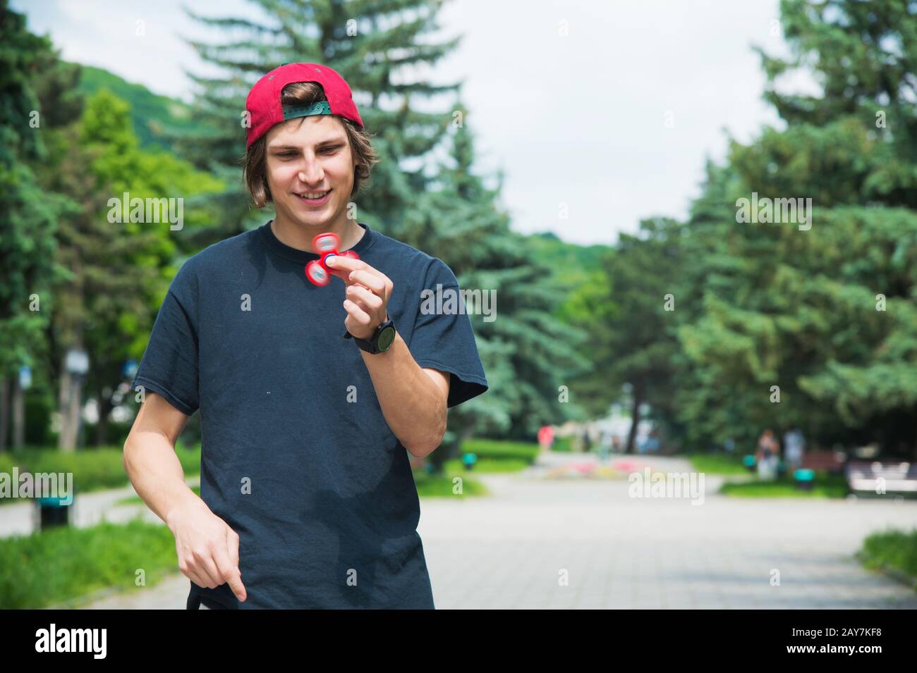 A long-haired hipster in a cap sits on a bench and spins a fidget-spinner Stock Photo