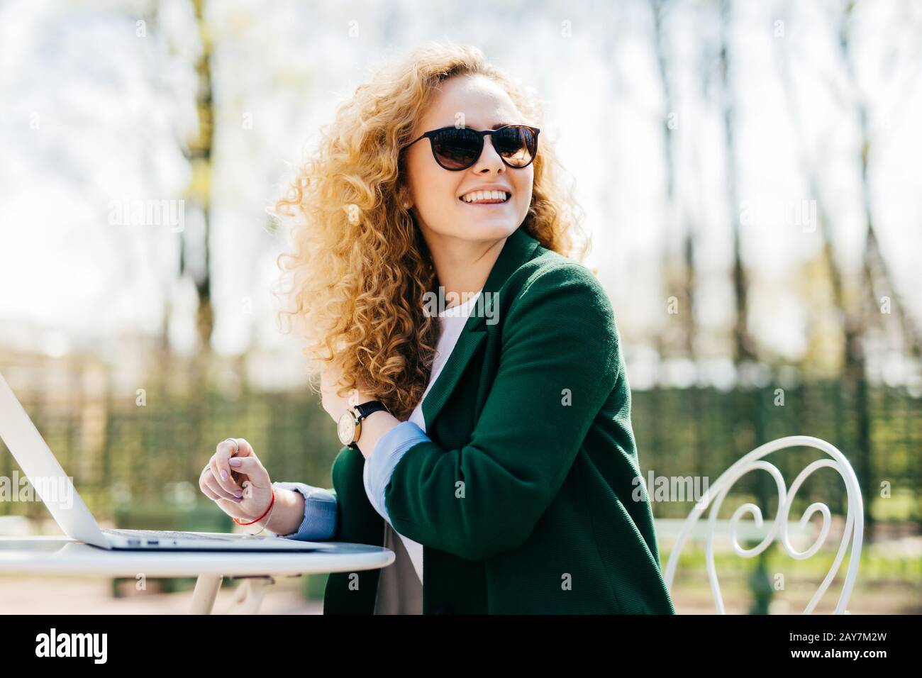 Happy stylish woman with curly light hair wearing sunglasses working with laptop outside in park typing necessary documents turning back noticing some Stock Photo