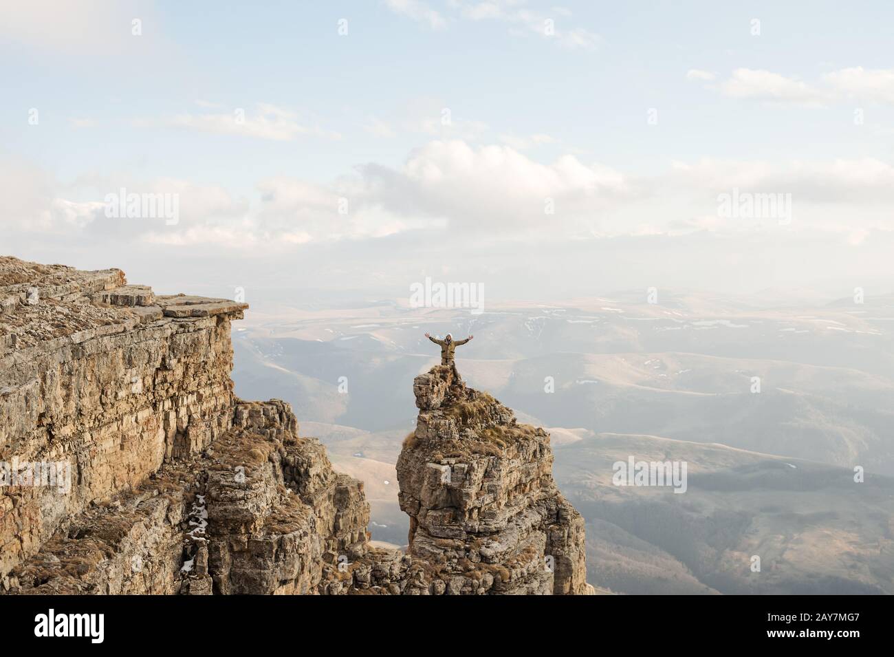 A happy man with his hands up high stands on top of a separately standing rock that is above the clouds against the background o Stock Photo