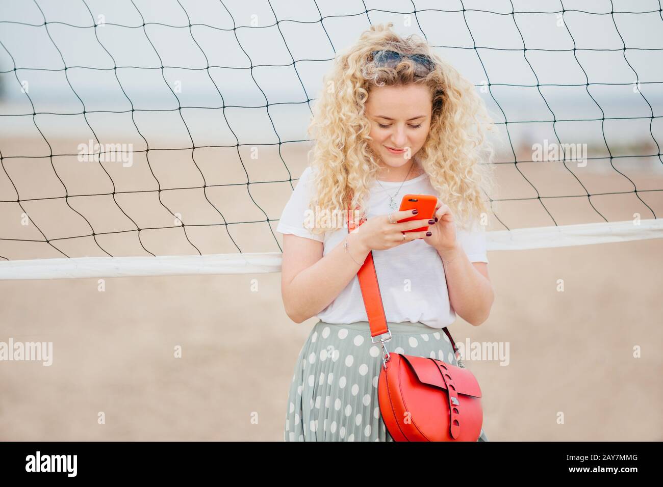 Oudoor shot of pleasant looking blonde female holds cell phone, shares photos in social networks, dressed casually, uses free internet, has walk acros Stock Photo