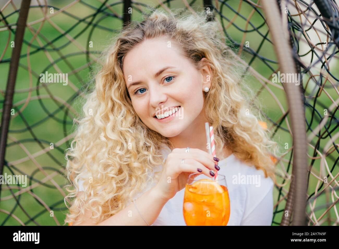 Outside shot of good looking curly female with friendy exprression, drinks summer fresh cocktail, poses in hanging chair, has positive smile, breathes Stock Photo