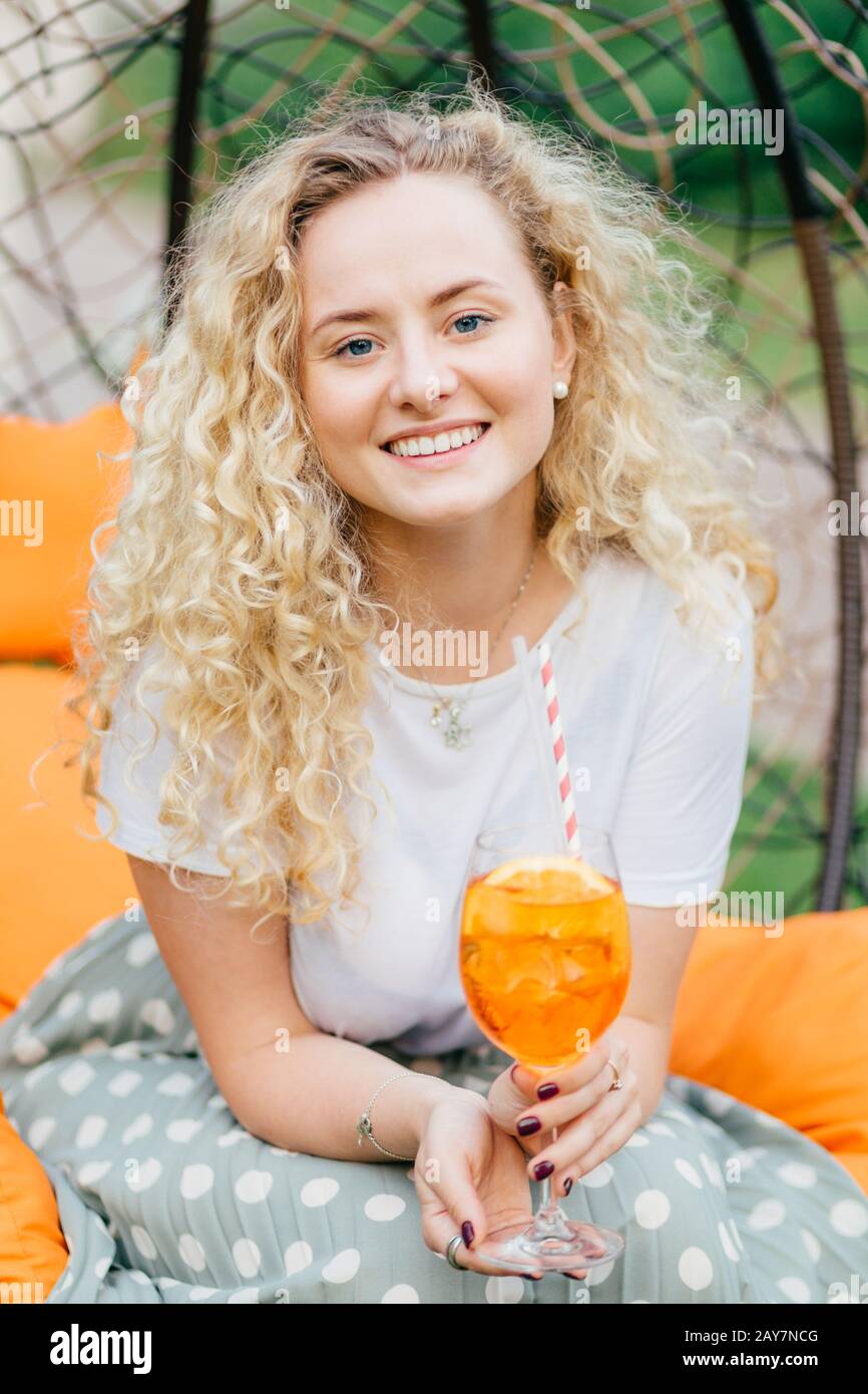 Attractive young female with charming smile, has wavy hair, dressed casually, holds fresh summer cocktail, recreats in garden during weekend. People, Stock Photo