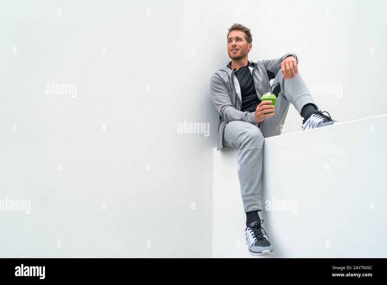 Healthy sport man drinking green smoothie during training at gym in activewear sweatpants outdoor. Athlete sitting relaxing post-workout. Stock Photo