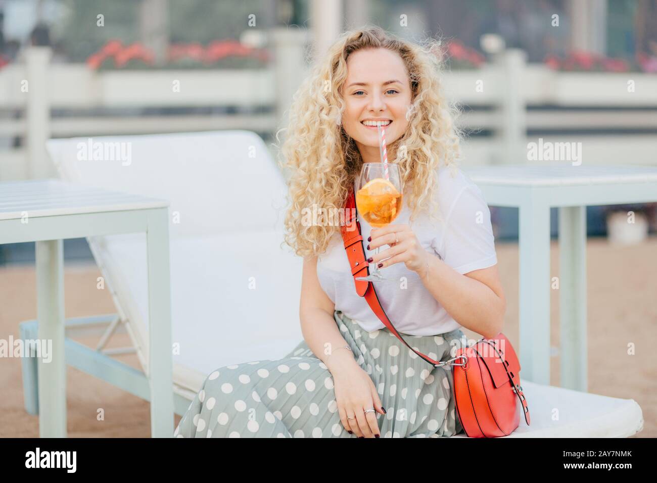 Happy blonde woman with curly light hair, holds fresh orange cocktail, being in high spirit, dressed casually, poses outdoor on beach resort, expresse Stock Photo