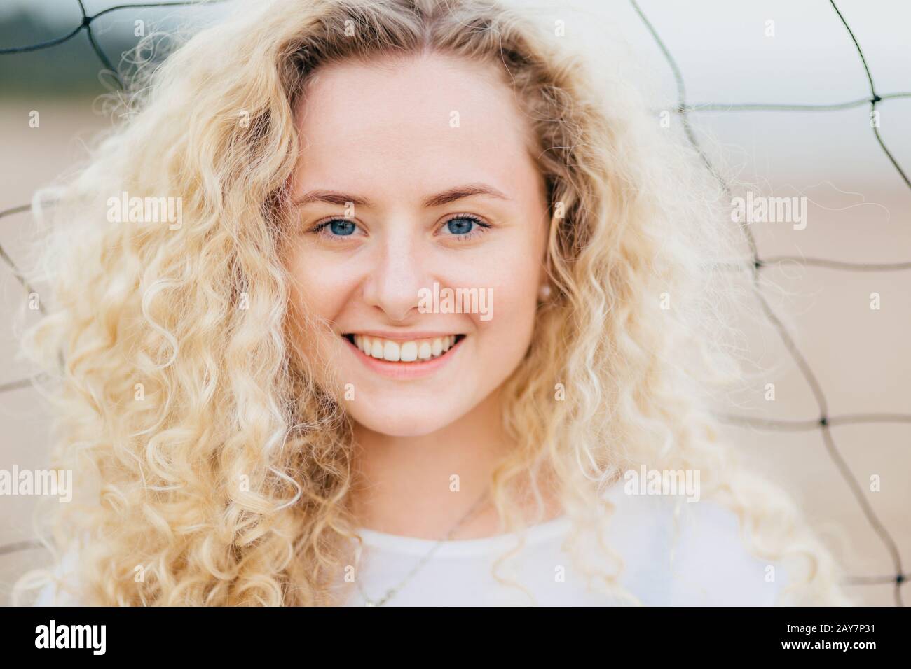 Glad curly Caucasian woman with toothy smile, has curly bushy light hair, blue eyes, clean skin, stands outdoor, has appealing appearance. Close up po Stock Photo