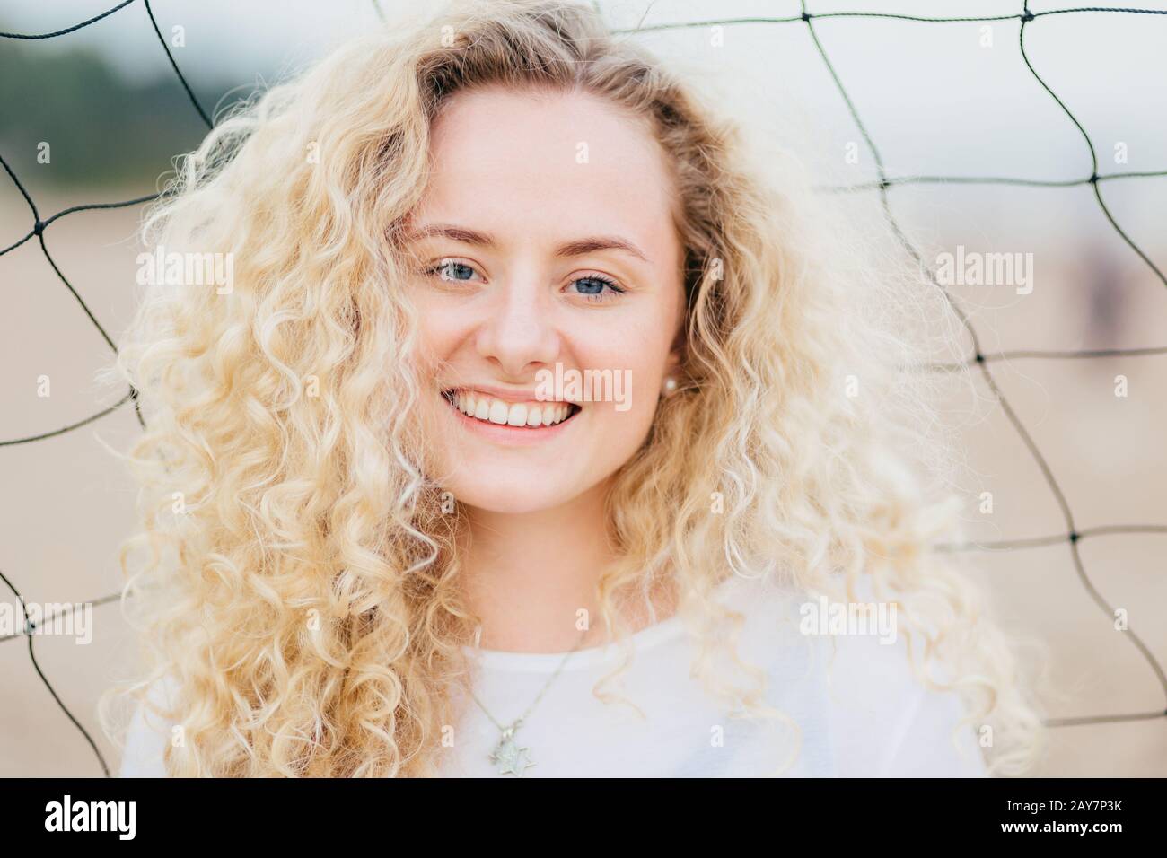 Lovely cute young woman has fresh skin, curly light hair, smiles gladfully, dressed in casual clothes, stands near tennis net, being satisfied to reci Stock Photo