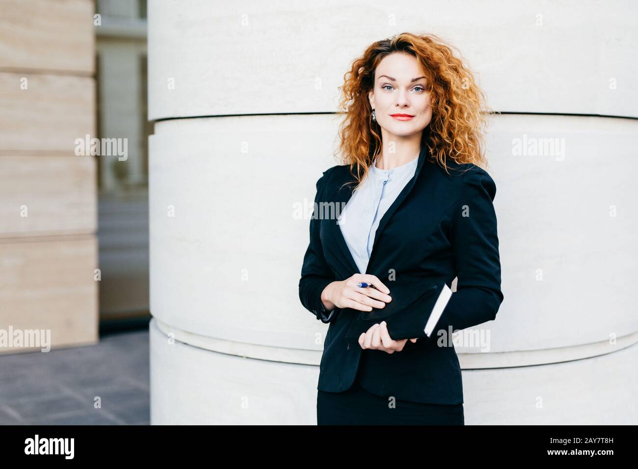 Businesswoman with curly light hair, red painted lips, wearing white blouse, black jacket and skirt, holding notebook with pen, going to write necessa Stock Photo