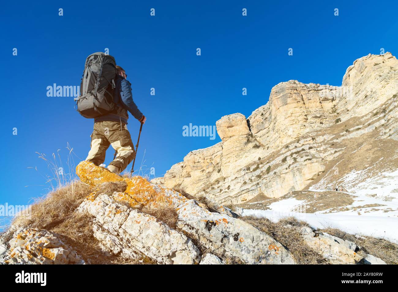 Backpacker with a large backpack and sticks ascends to the rock against the background of epic rocks in the winter season. Stock Photo