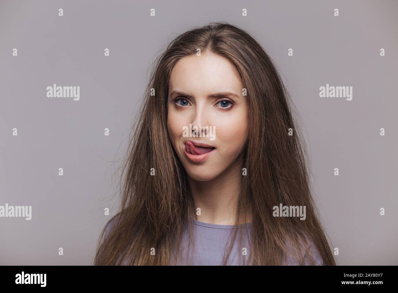 Shot of beautiful brunette female licks lips with tongue, has pure healthy skin, dressed casually, isolated over grey background. Lovely woman shows t Stock Photo