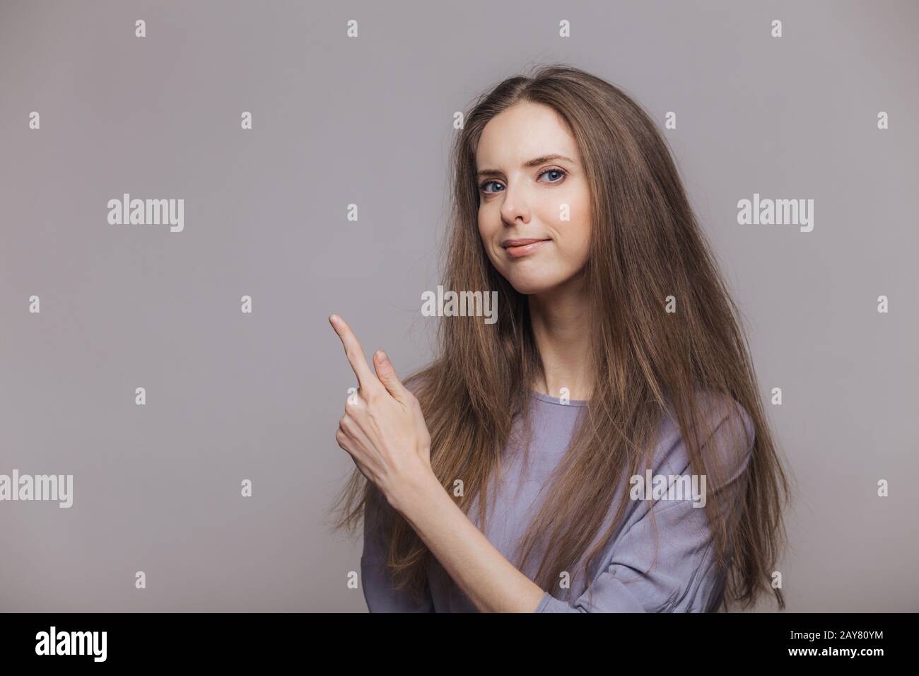 Shot of serious pleasant looking brunette female with blue eyes, dressed casually, points with index finger at blank copy space, isolated over grey ba Stock Photo