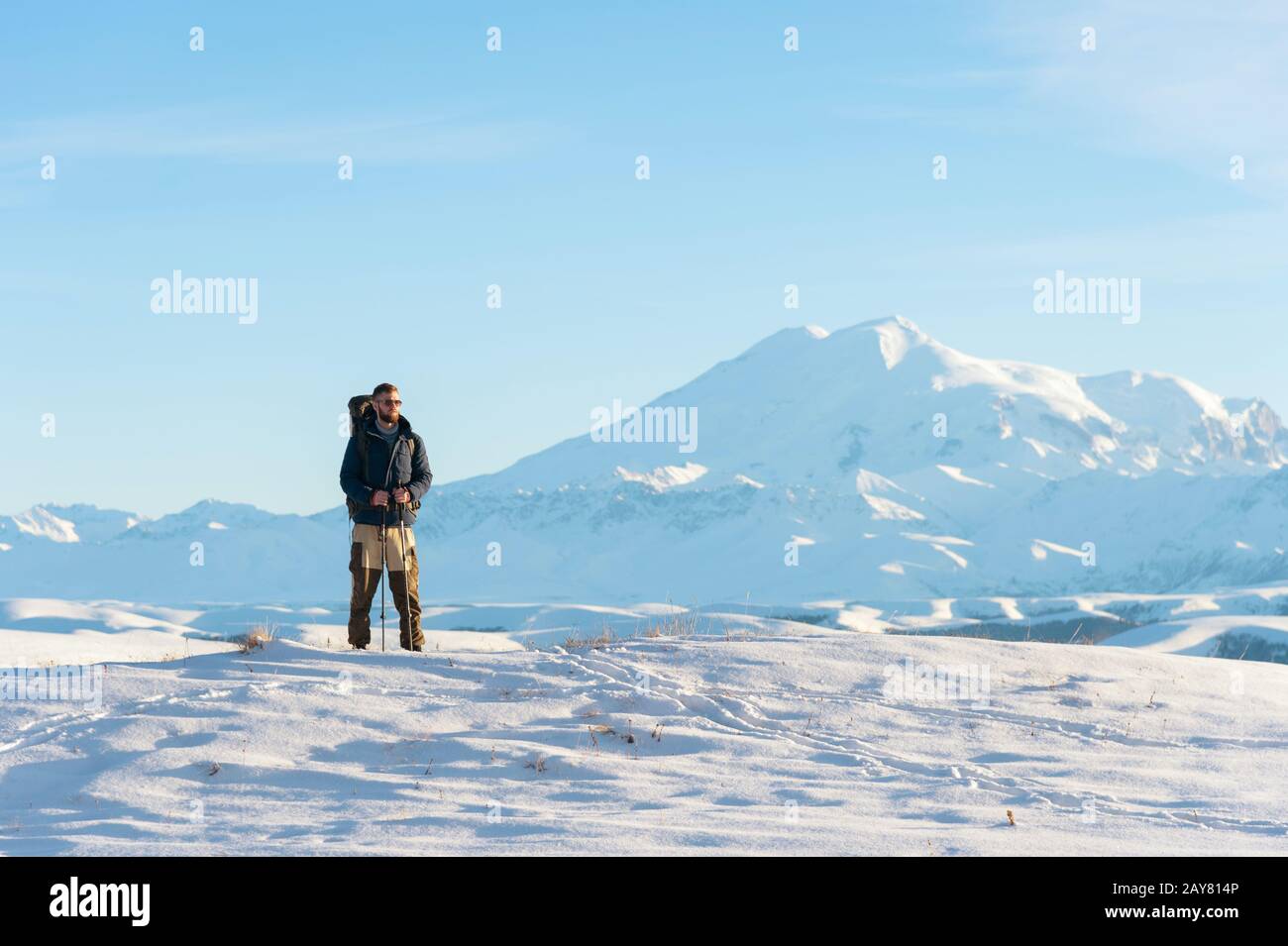 A traveler with a large backpack on his shoulders Stand on a snow-capped hill against the blue sky and the sleeping Elbrus volca Stock Photo