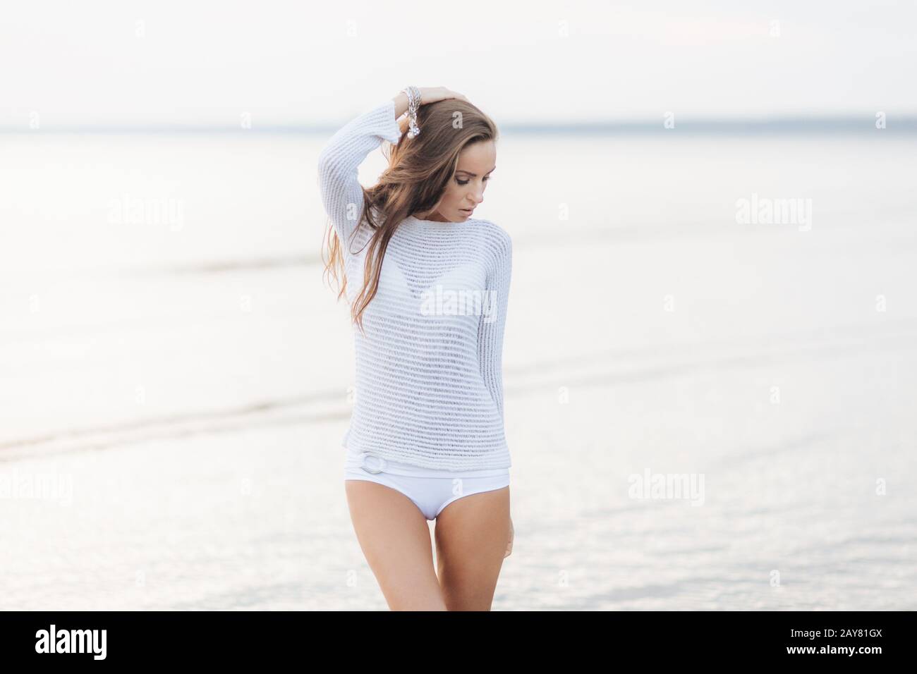 Adorable young female model in white sweater and bikini, stands against beautiful seascape, looks thoughtfully down, dreams about something pleasant a Stock Photo