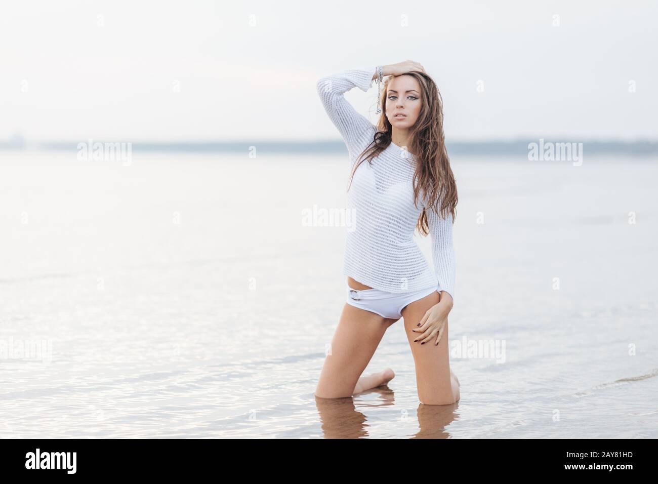 Outdooor view of good looking brunette female with make up, dressed in white bikini, poses at camera near calm sea background, has fit body shape, fee Stock Photo