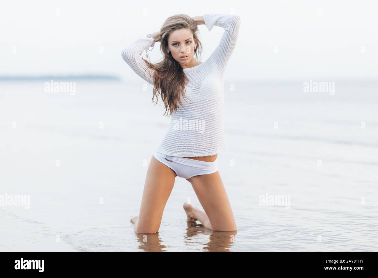 Portrait of attractive female with make up, dressed in white clothes, stands on knees at sandy beach near ocean view, has confident expression, feeles Stock Photo