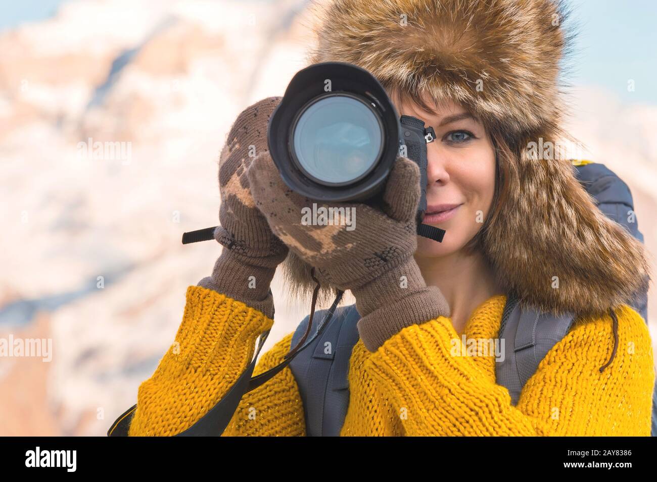 Portrait of a sweet tourist girl in a big fur hat takes pictures on her digital camera in the mountains. Stock Photo