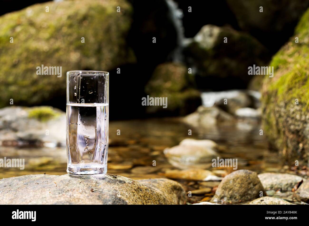 A transparent glass glass with mineral mountain river water stands on a stone beside the mountain river creek Stock Photo
