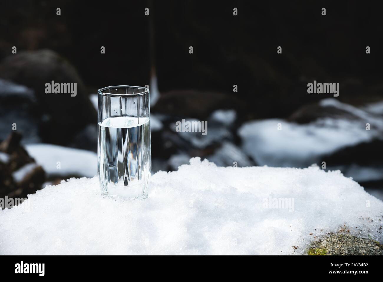 A transparent glass glass with drinking mountain water stands in the snow against a background of a clean frost mountain river i Stock Photo