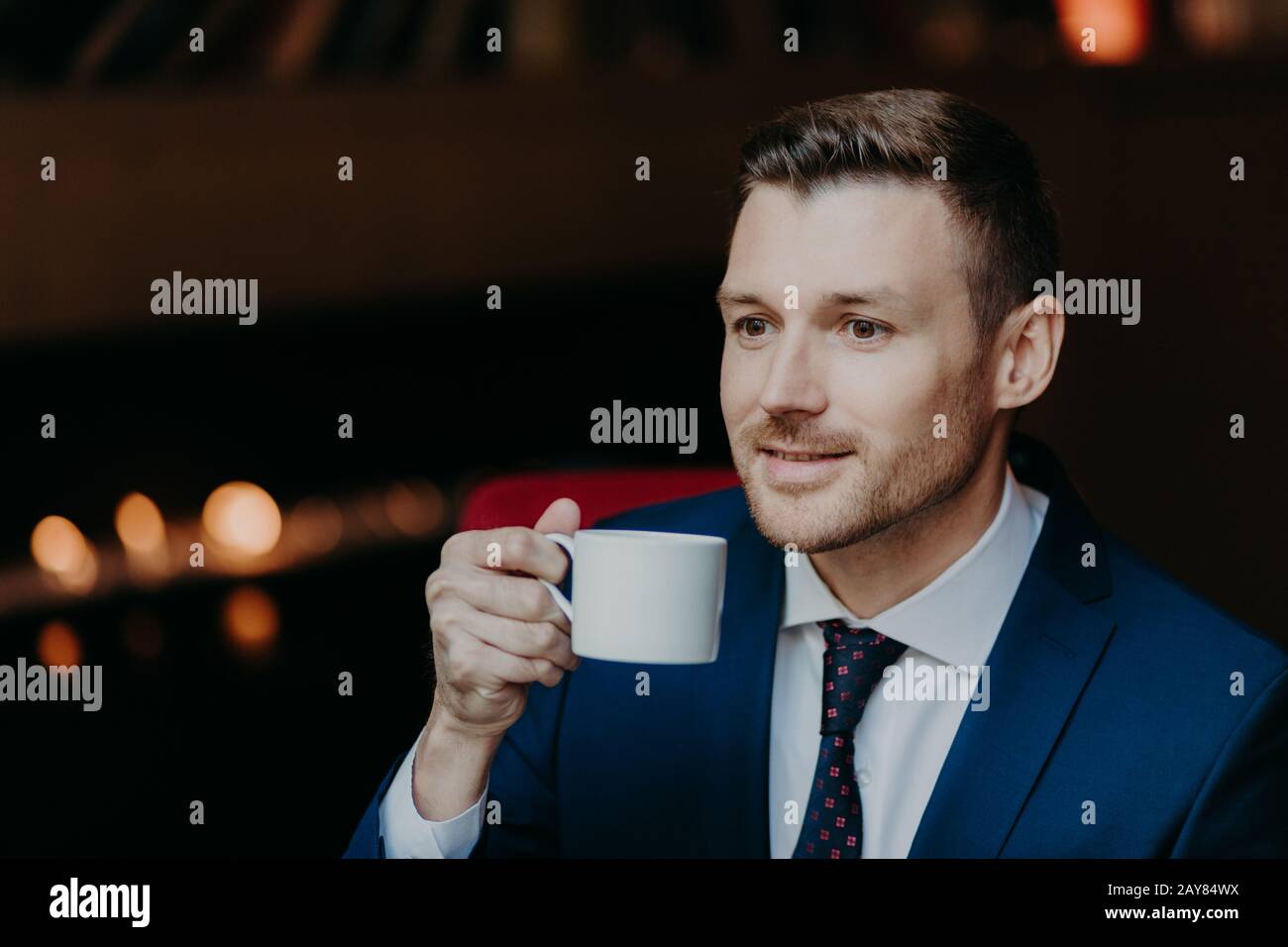 Thoughtful handsome young businessman in formal suit, holds cup of coffee, being deep in thoughts, thinks about new startup, recreats during dinner br Stock Photo