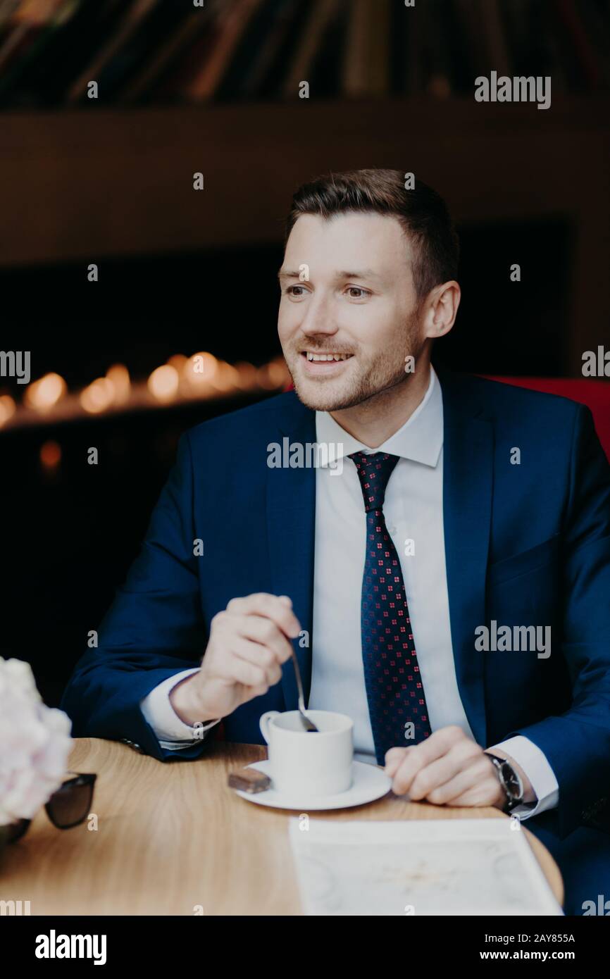 Vertical shot of handsome male in formal clothing, has cheerful expression, has dinner break at cafeteria, drinks aromatic coffee, looks happily somew Stock Photo