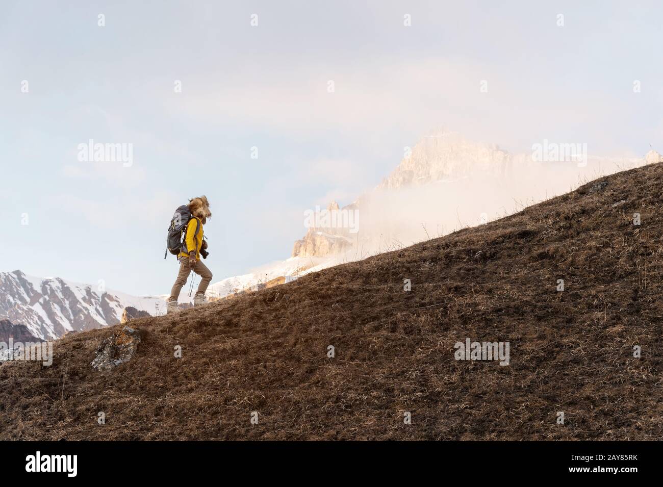 A backpacker in a big fur hat and gloves with a backpack on his back goes uphill against the background of epic cliffs in the cl Stock Photo
