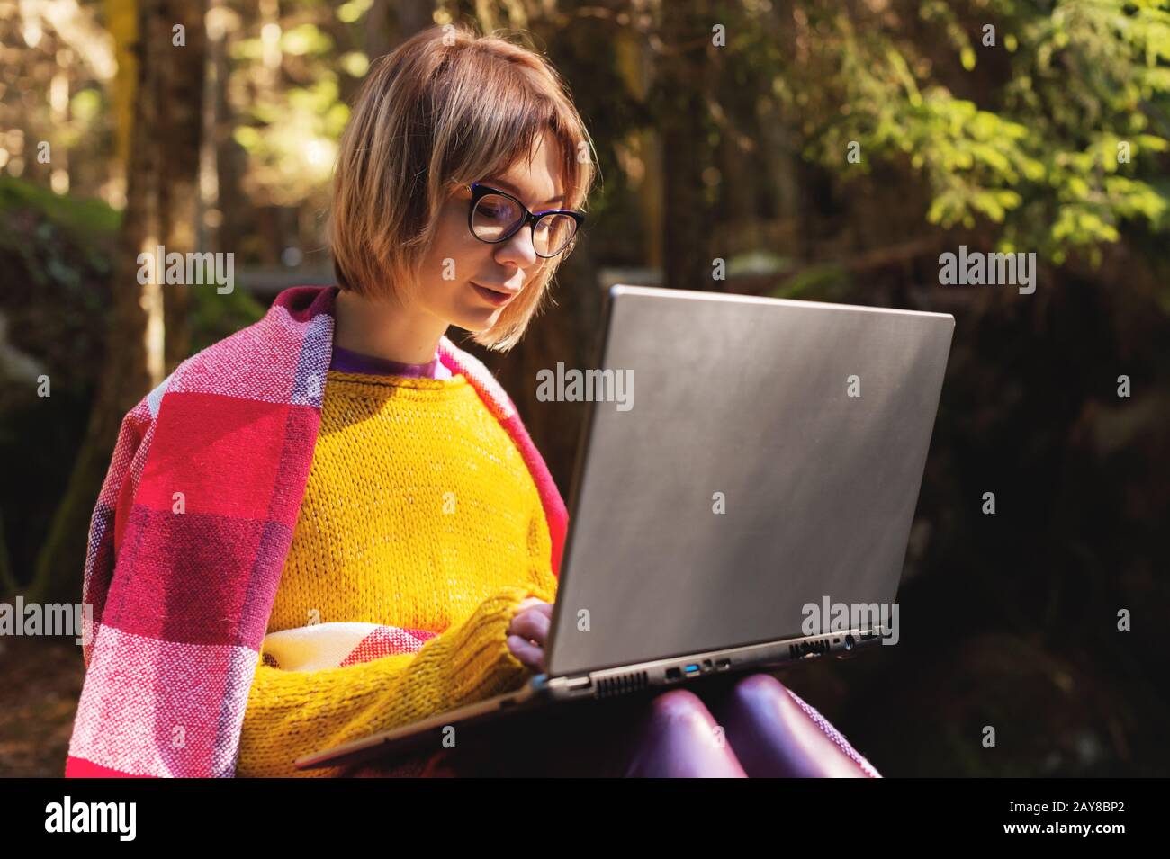 A toned portrait of a a smiling freelancer hipster girl with glasses dressed in a blanket with a laptop kneeling sitting on a ro Stock Photo