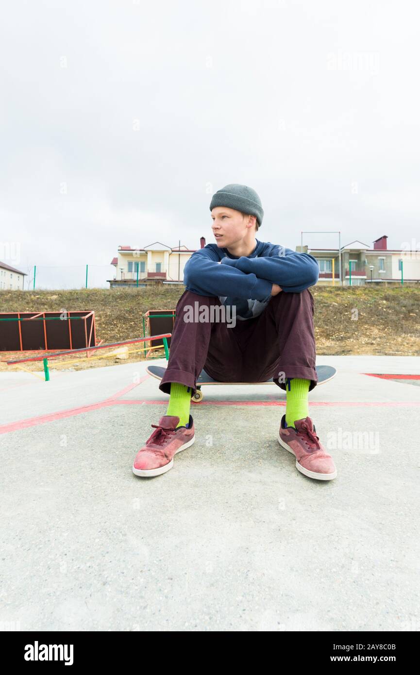 A teenage boy is sitting on a skateboard in the park. The concept of free time pastime for teenagers in the city Stock Photo