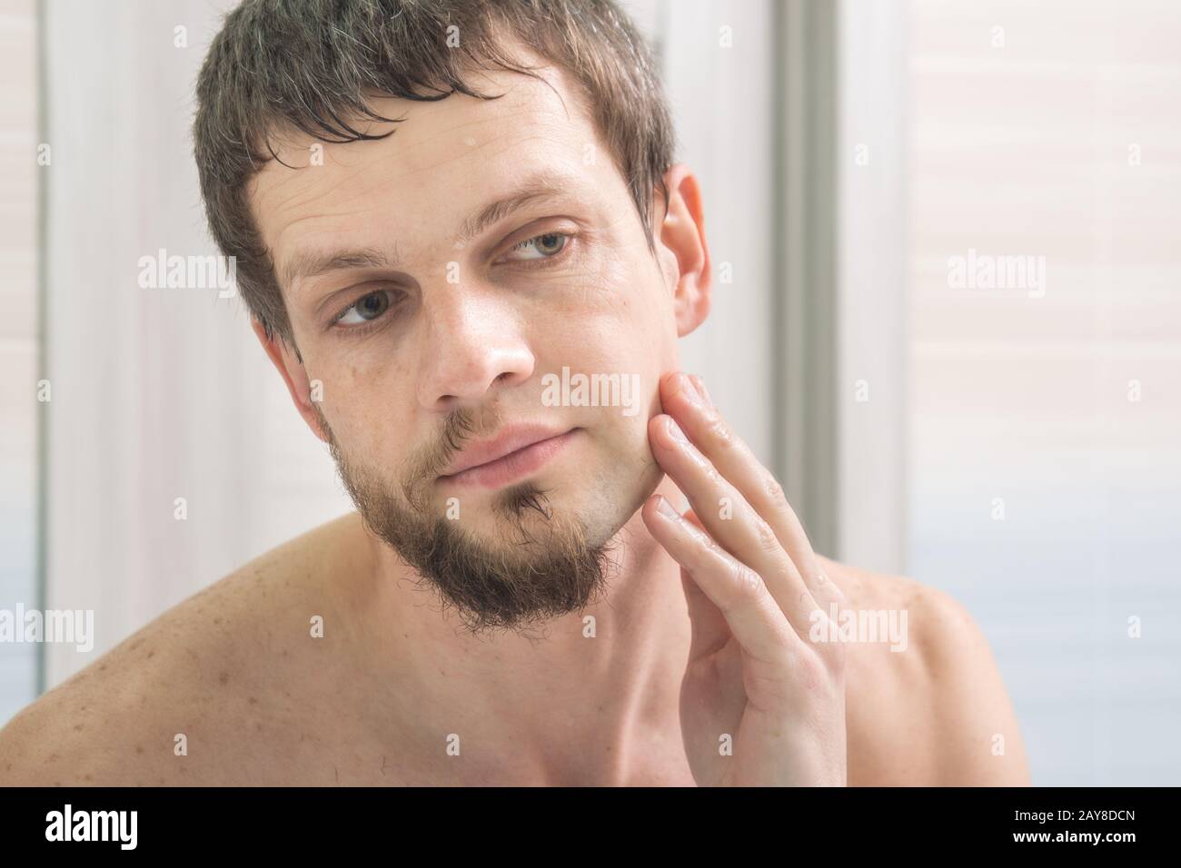The guy shaved one half of his face and looks in the mirror evaluating the result Stock Photo