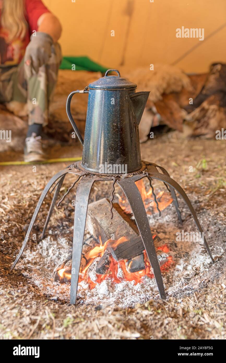Old metal coffee pot stands over a campfire on a rack. Stock Photo