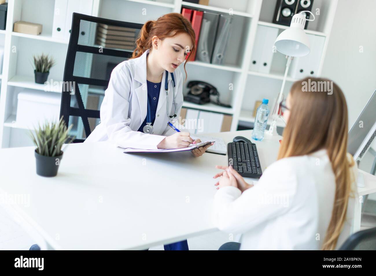 A beautiful young girl in a white robe is sitting at the desk in the office and communicating with the interlocutor. The girl ma Stock Photo