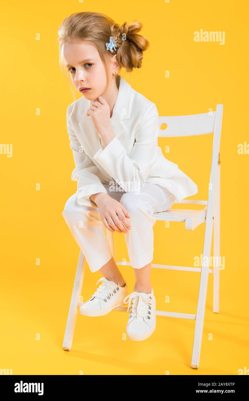 A teenage girl in white clothes is sitting on a chair on a yellow background. Stock Photo