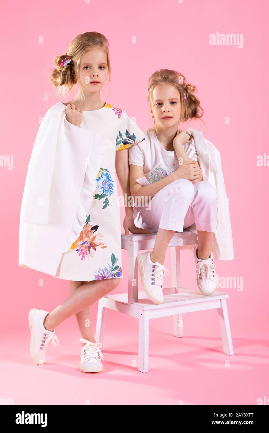 Twins girls in bright clothes posing near the stairs with two steps on a pink background. Stock Photo