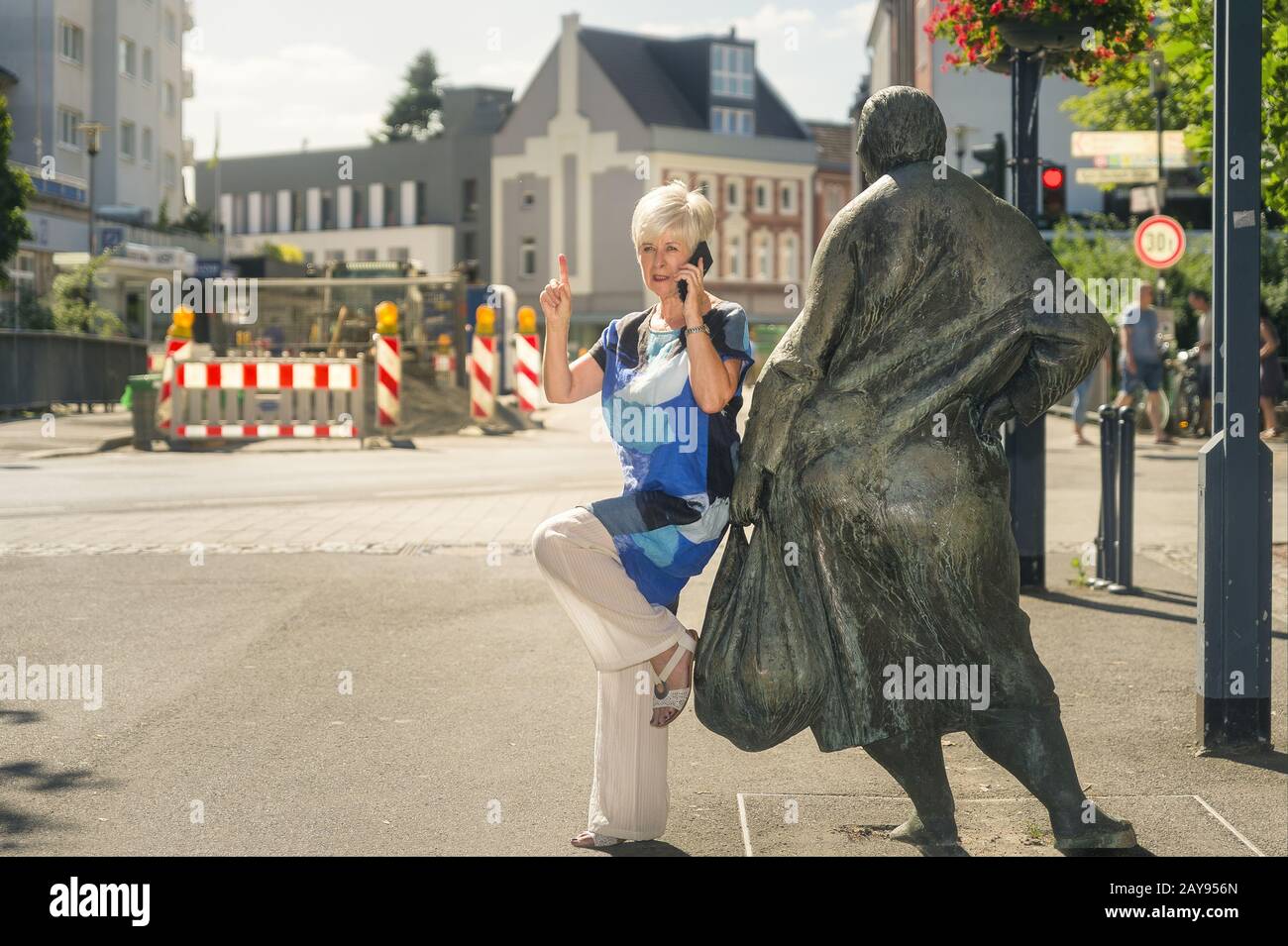 A senior citizen leans against a bronze figure and calls on a smartphone. Stock Photo