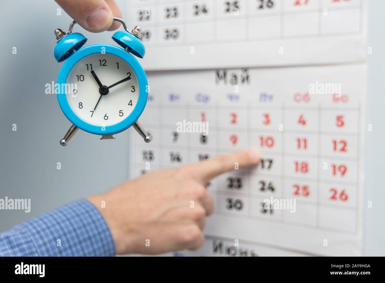 A hand holds a small alarm clock, in the background a hand points to long weekends and holidays on a wall calendar Stock Photo