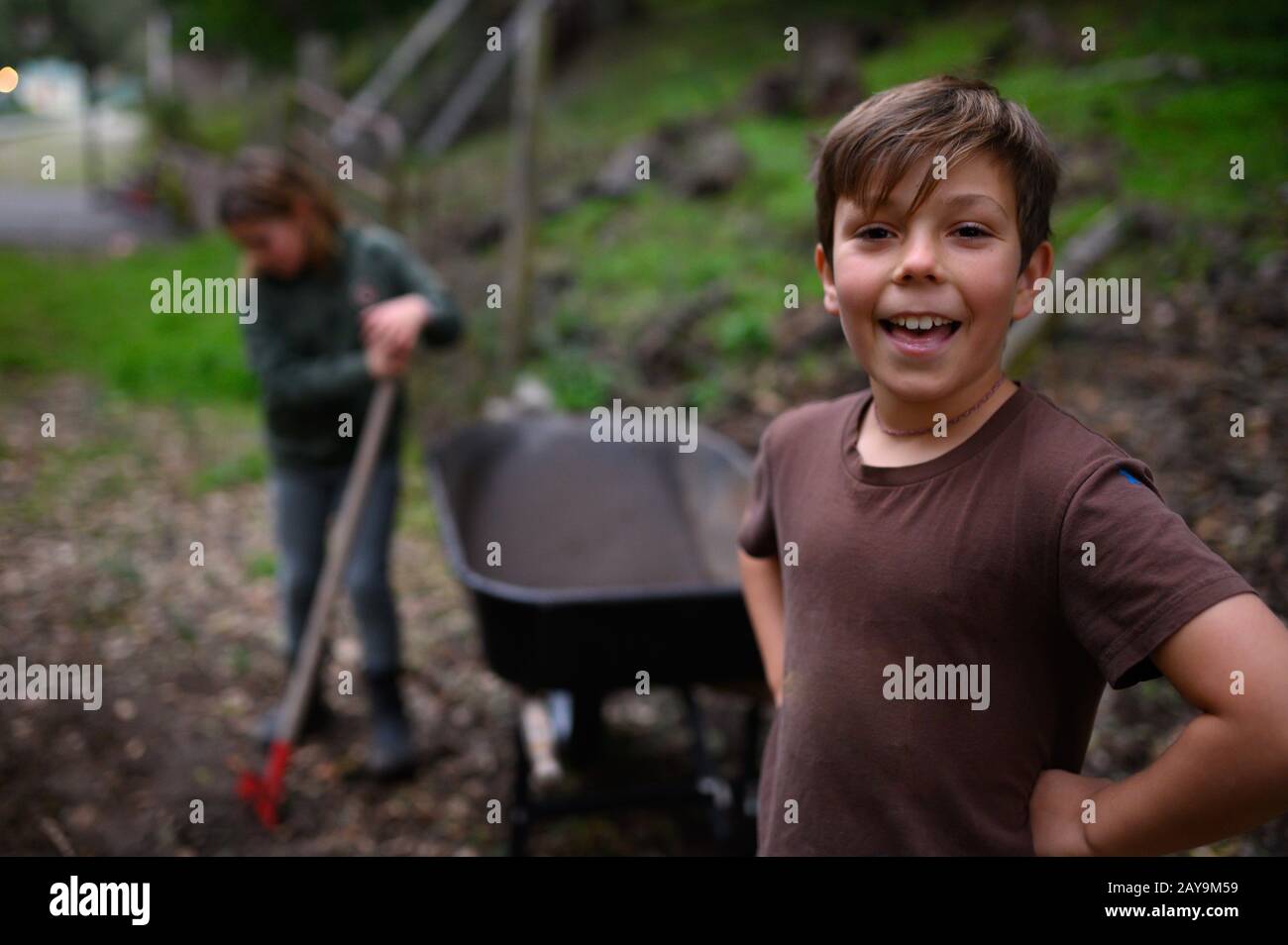 Smiling boy in a brown t-shirt standing near wheelbarrow in yard Stock Photo