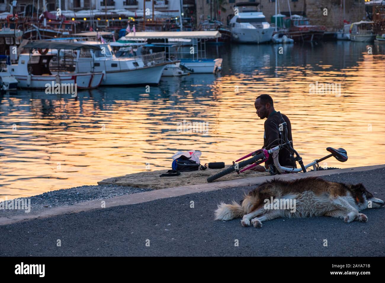 An african man stands by the sea at the old harbor of Kyrenia in Cyprus whilst his dog is sleeping on the tarmac Stock Photo