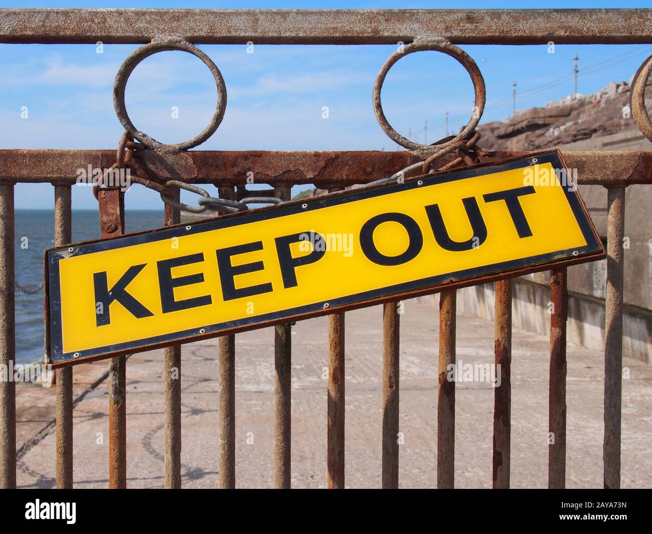 yellow keep out sign on a rusty metal closed across a road leading to the sea Stock Photo