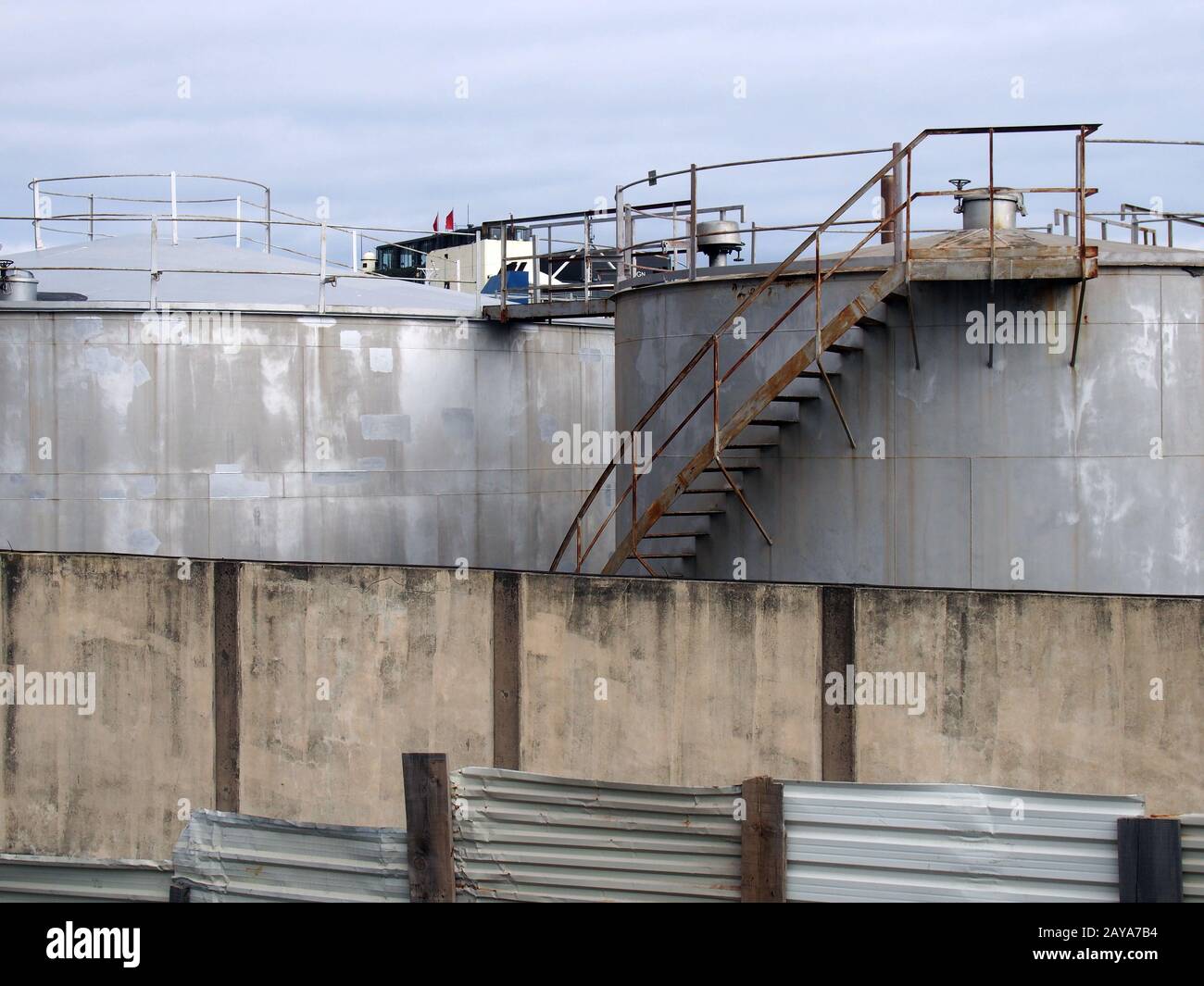 old steel industrial storage tanks with rusty stairs and walkways behind a shabby corrugated iron fence and wall Stock Photo