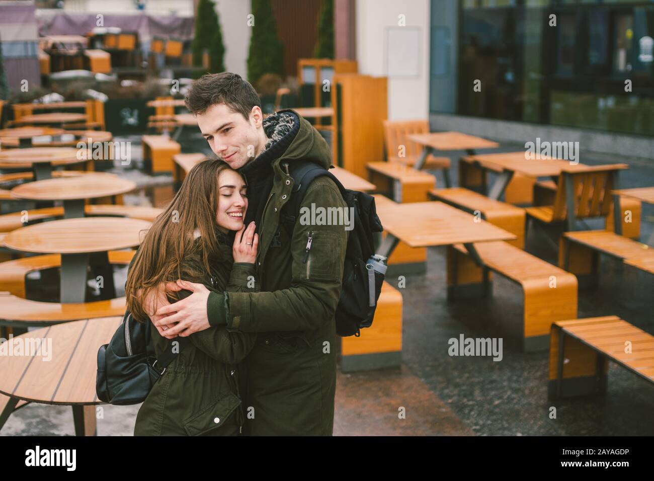 A young couple in love and a girl and a student stand embracing near the tables of a street terrace cafe closed empty without pe Stock Photo