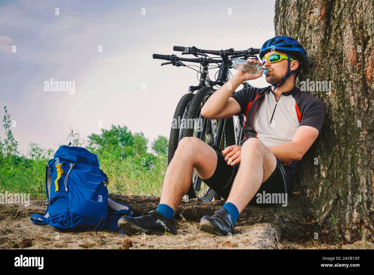 Caucasian male cyclist active lifestyle resting under tree after exercising bicycle drinking water from bottle. Sportsman in hel Stock Photo