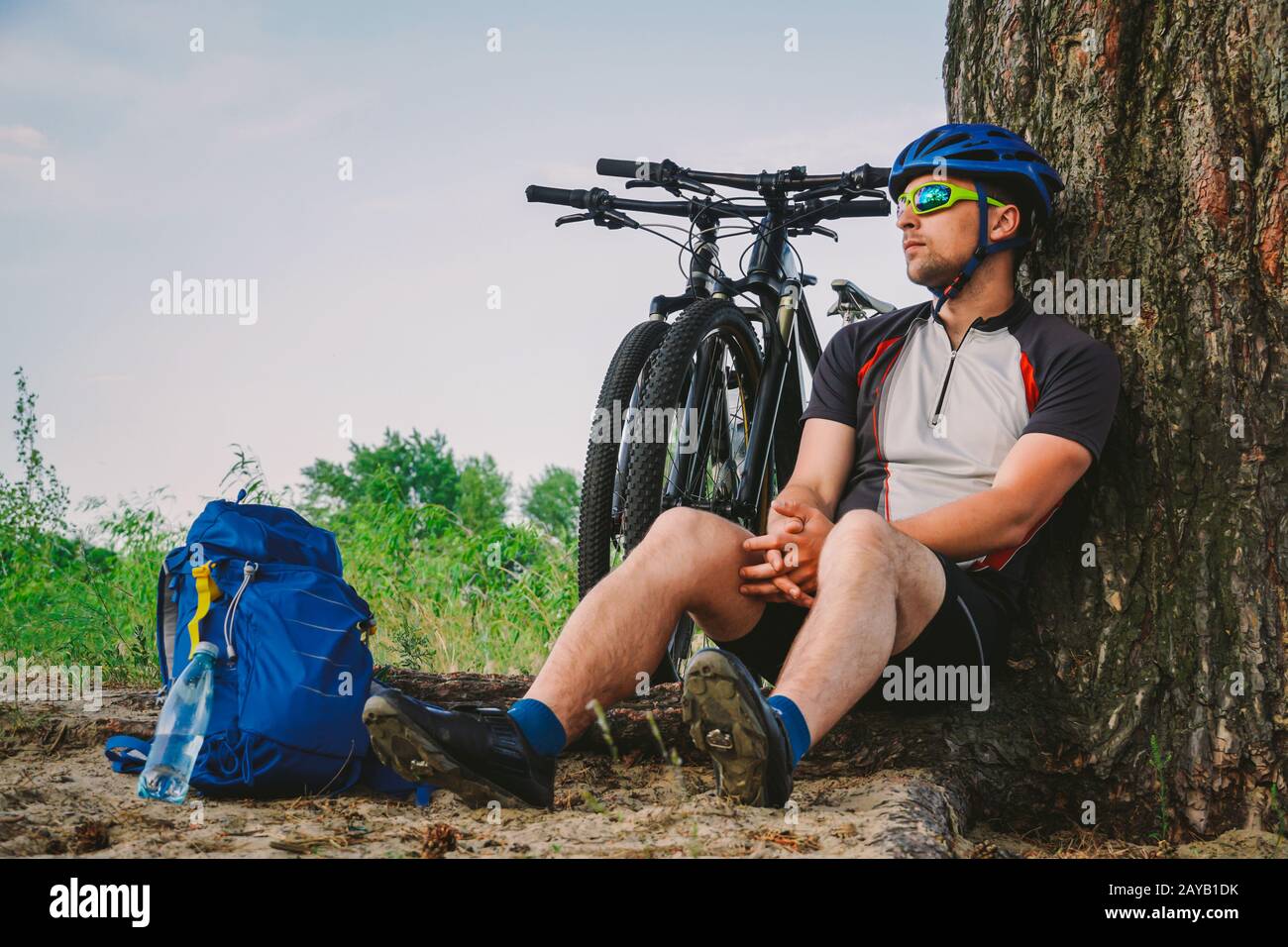 male mountain biker resting on bike ride, sitting on ground under a tree with his mountain bike, stands next to him, enjoying th Stock Photo
