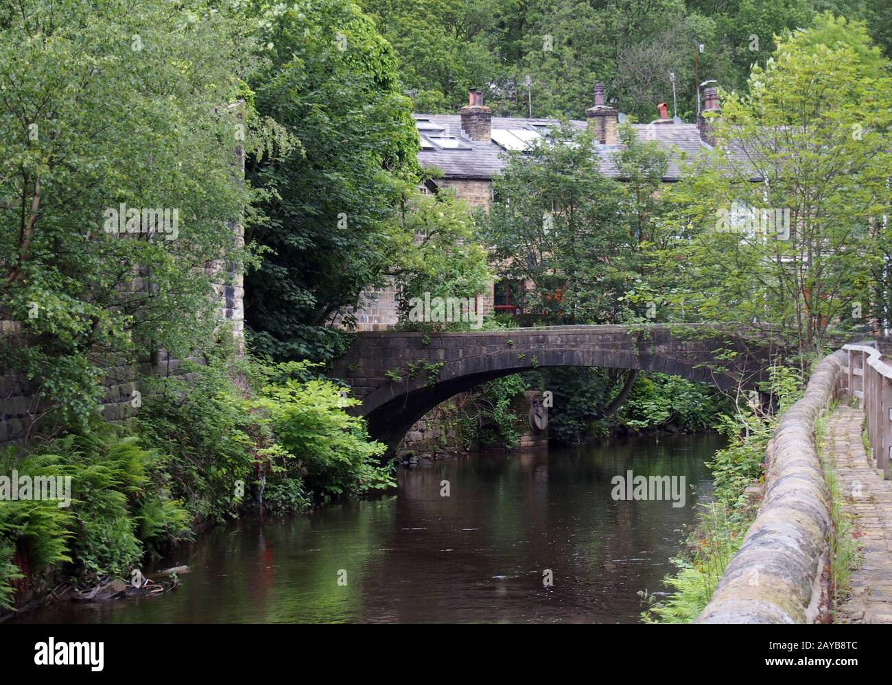 a footbridge crossing the river calder at eastwood near hebden bridge with a row of stone houses amongst forest trees Stock Photo