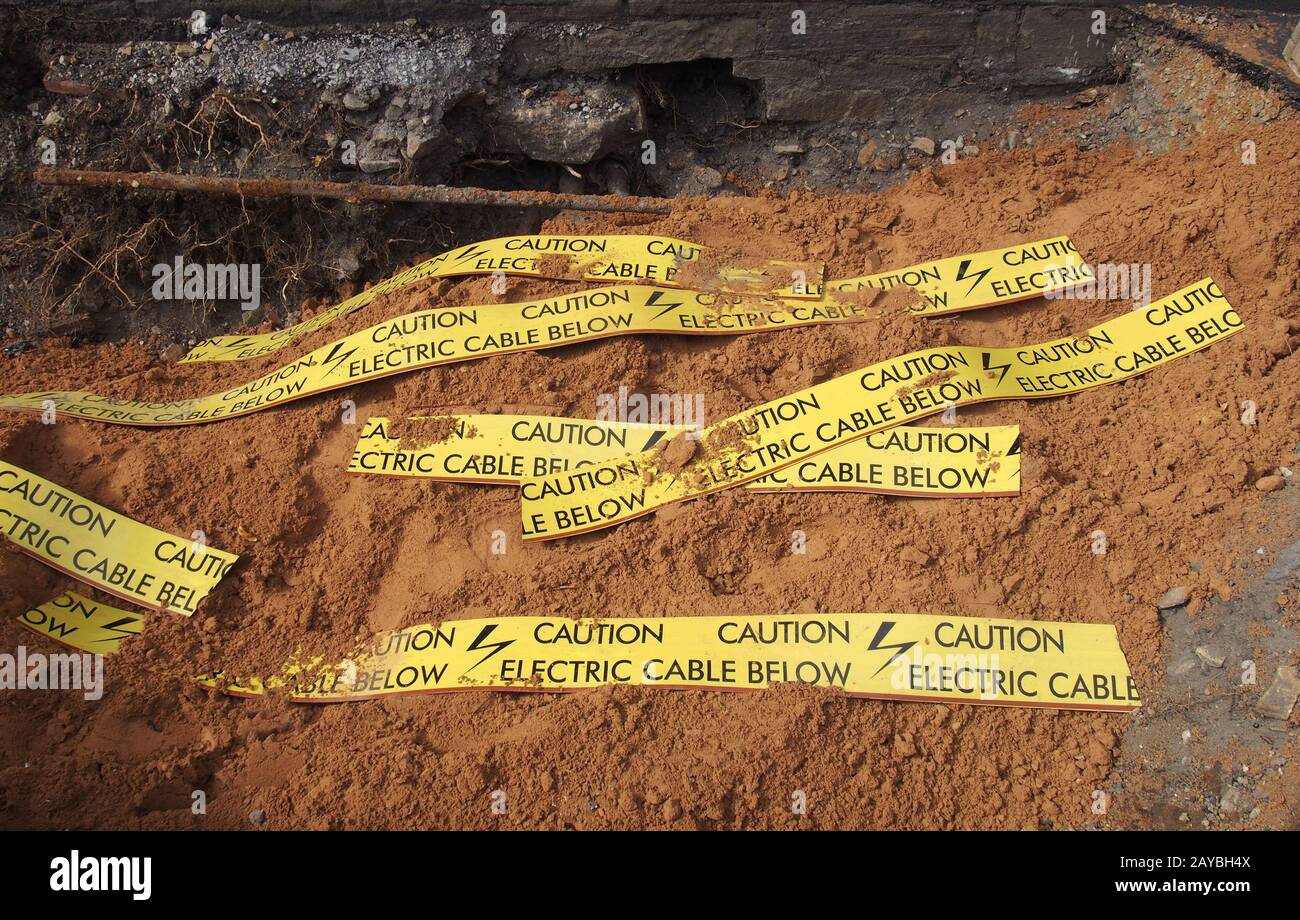 yellow tape signs warning of buried electric cable below in a trench being excavated during roadwork and building construction Stock Photo