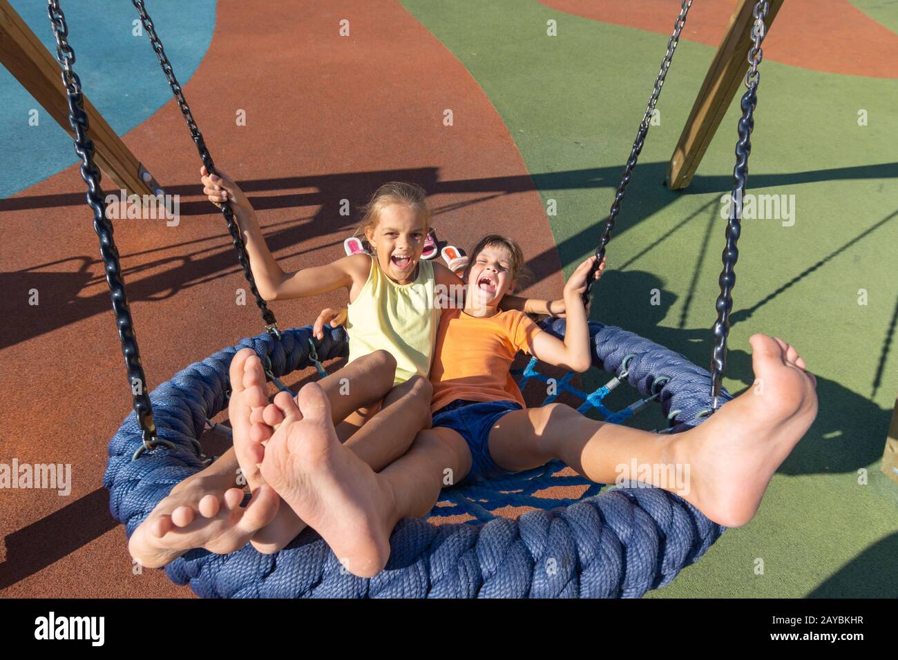 Two girls joyfully and cheerfully ride on a large hanging swing in the playground Stock Photo