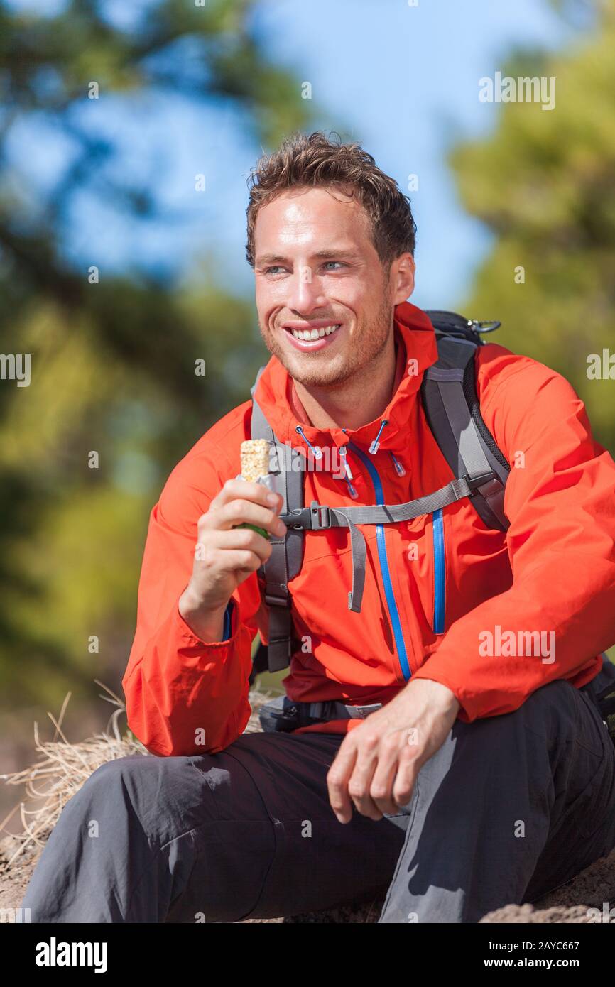 Hiker man healthy outdoor lifestyle eating muesli bar during hike on mountain hiking. Happy people eat granola cereal bar snack living active lifestyle in nature. Stock Photo