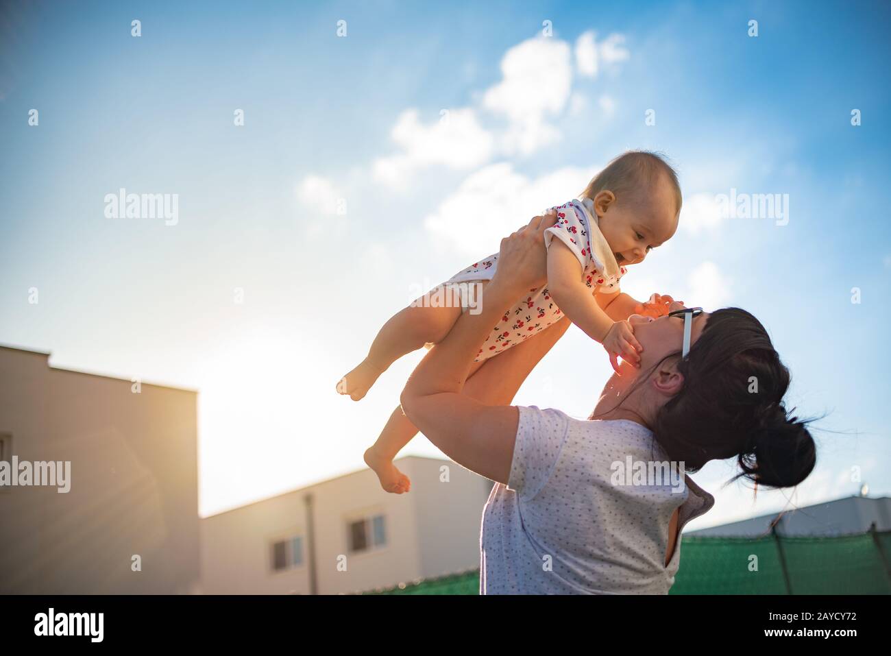 An emotional picture of 1 year old baby and her mother holding her up in the air against blue sky and bright sun Stock Photo