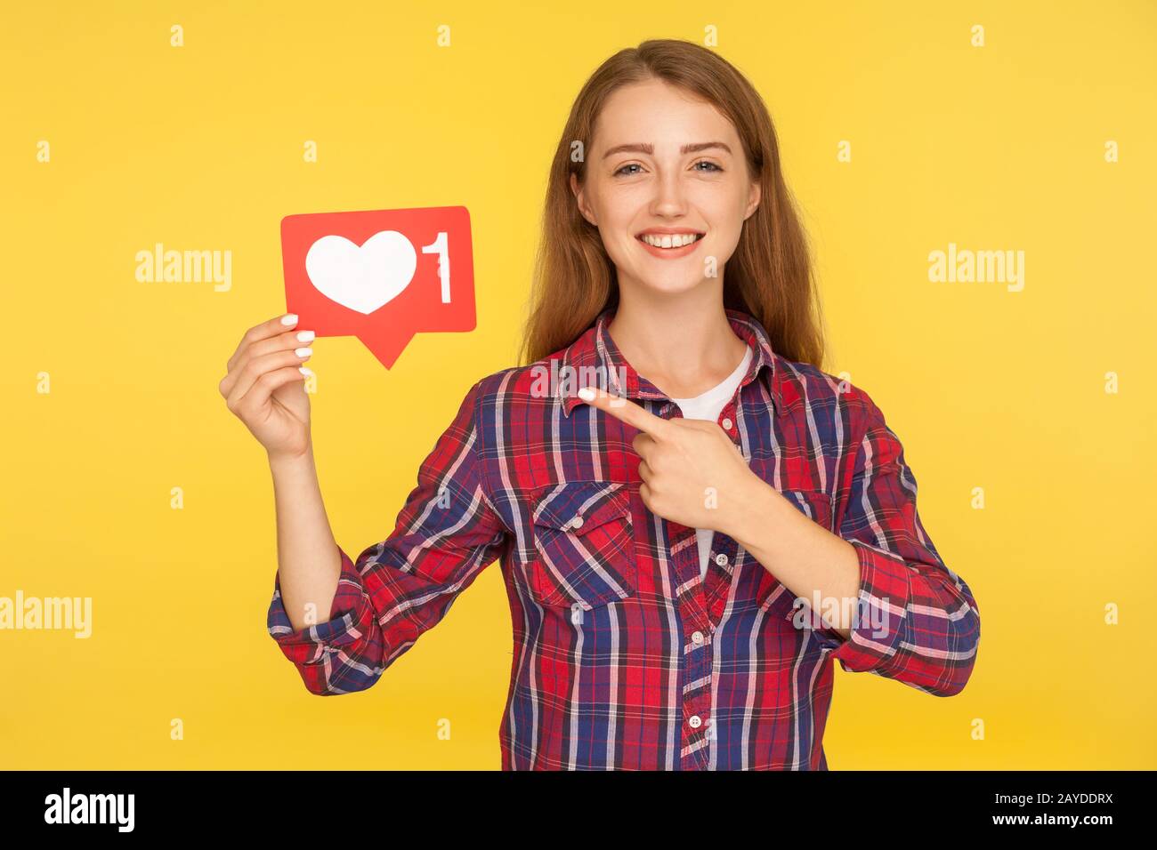 Push heart button to enjoy content. Portrait of happy ginger girl in checkered shirt pointing at social media like icon, follower notification symbol. Stock Photo