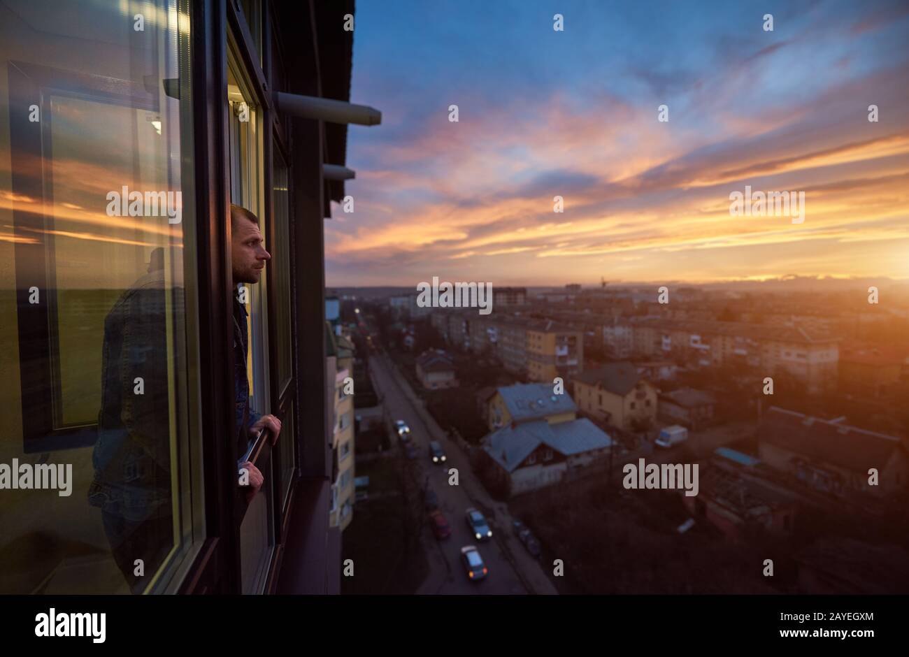 Adult man stands on the balcony and looks into the distance enjoying the sunset. Beautiful city landscape of a fiery sky at sunset. Roofs of houses and passing cars in the bustle of the city Stock Photo