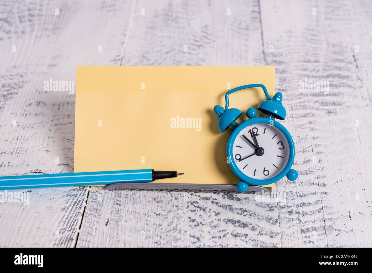 Mini blue colour alarm clock to show time placed tilted in front of one yellow notepaper. Classic little watch standing above a Stock Photo