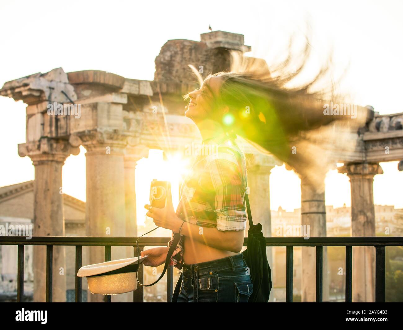 Beautiful young woman shaking long blond hair at Roman Forum at sunrise. Happy smiling tourist with vintage camera. Lens flair. Rome, Italy Stock Photo