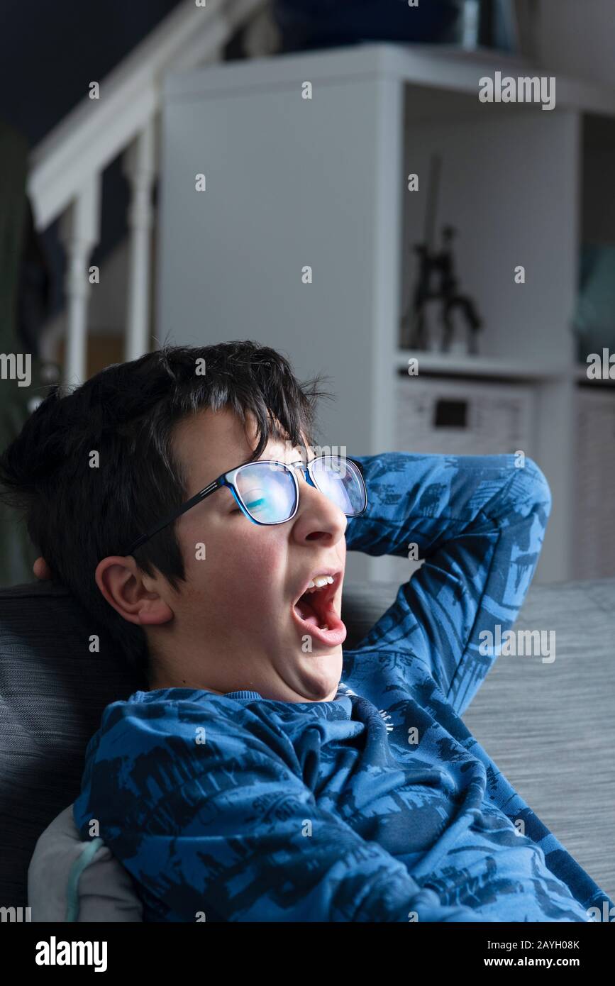 Bespectacled young boy yawning on the sofa , watching late night TV Stock Photo